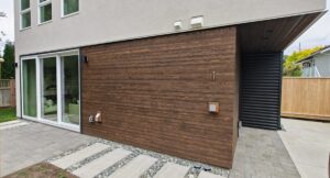Modern home's exterior featuring dark wood cladding, a large glass sliding door, and a light gray upper facade with windows above.