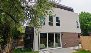 Modern two-story house with large glass doors and beige stucco facade, wood siding, and a wooden fence in a green backyard.