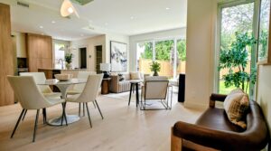 Open-concept living and dining area with white dining chairs around a round glass table, beige sofa, and large windows showing a green backyard and plants inside.