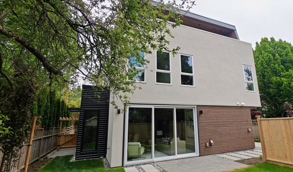 The URBAN NEST back yard - Innovate Design Collective Modern two-story house with large glass doors and beige stucco facade, wood siding, and a wooden fence in a green backyard.