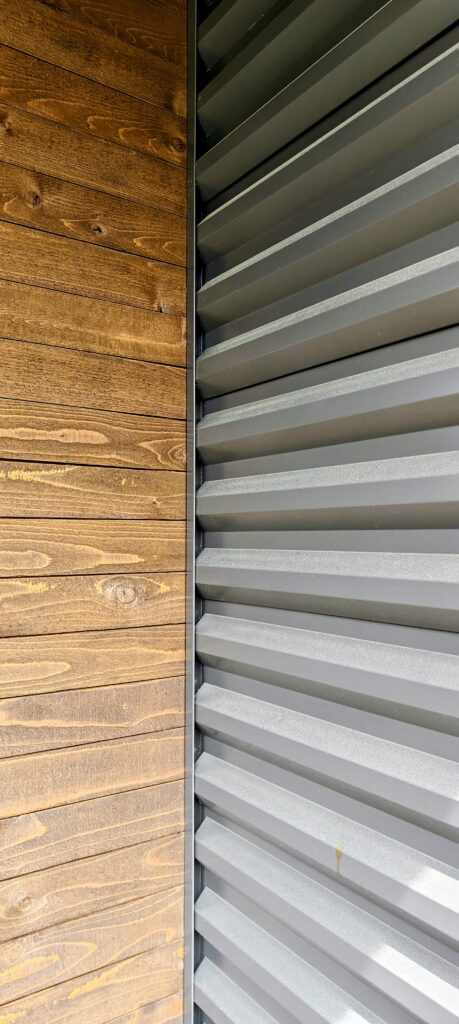 The URBAN NEST siding 2 - Innovate Design Collective Wooden wall paneling on the left with visible grain, meeting a right-side gray metal louvered surface at a sharp vertical seam.