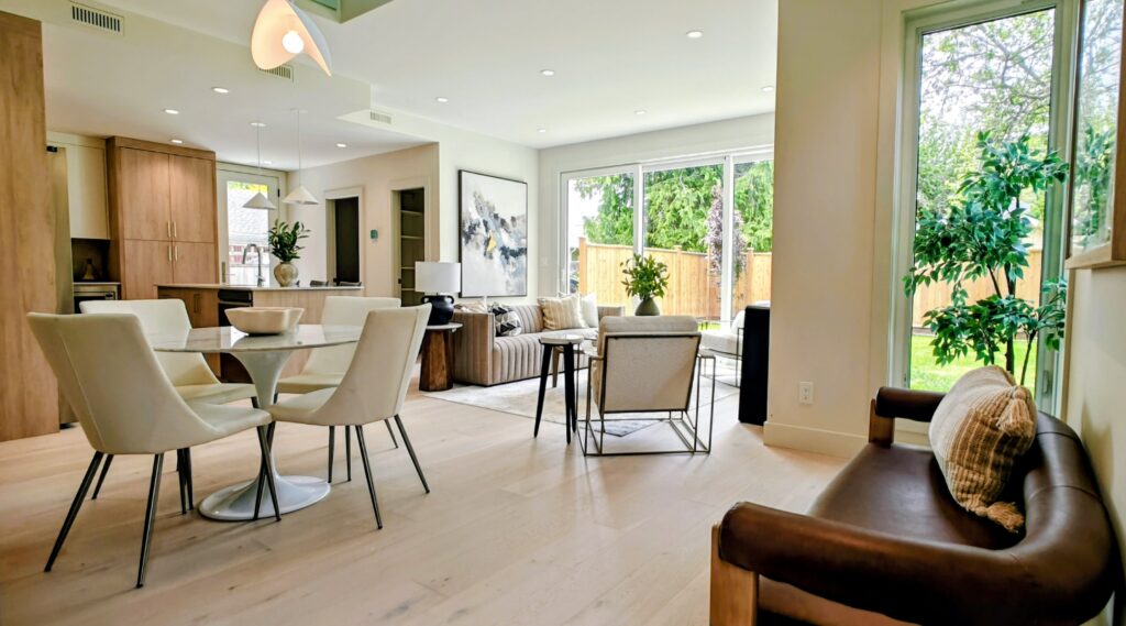 The URBAN NEST living 1 - Innovate Design Collective Open-concept living and dining area with white dining chairs around a round glass table, beige sofa, and large windows showing a green backyard and plants inside.