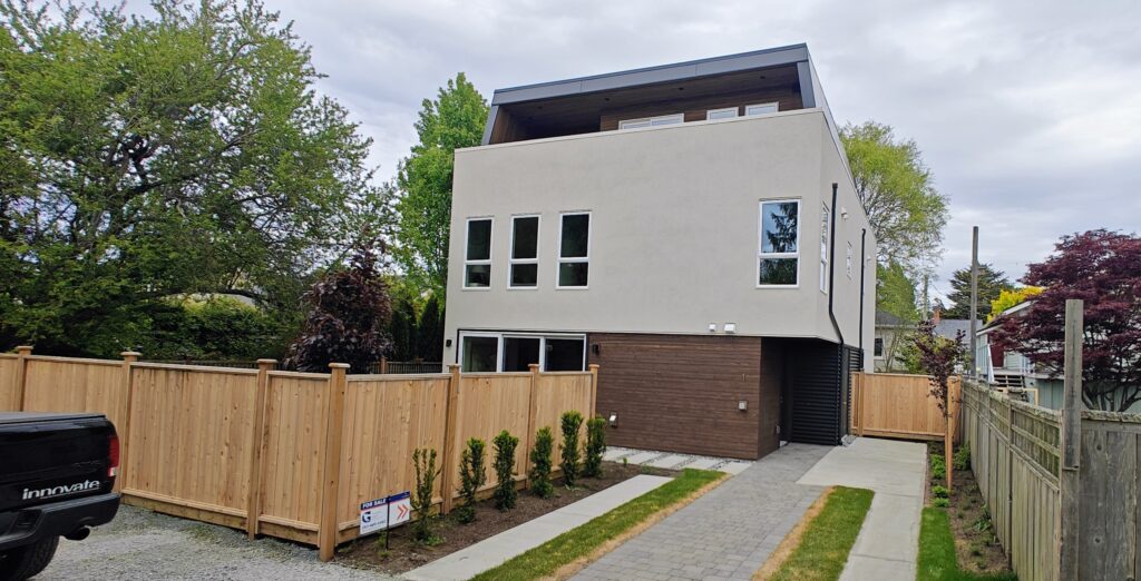 oplus_1048578 - Innovate Design Collective Modern two-story home with beige upper facade, dark brown lower siding, and a wooden fence along a paved driveway; trees in the yard.